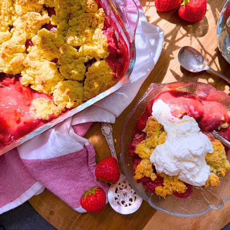 A baked strawberry dessert with a biscuit topping. There is a serving in front in a glass bowl. There is whipped cream on top.