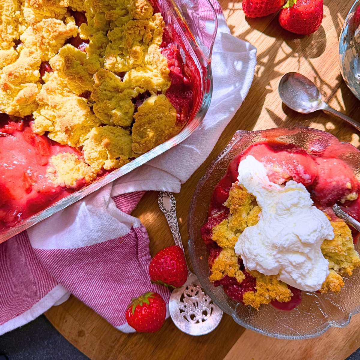 A baked strawberry dessert with a biscuit topping. There is a serving in front in a glass bowl. There is whipped cream on top.