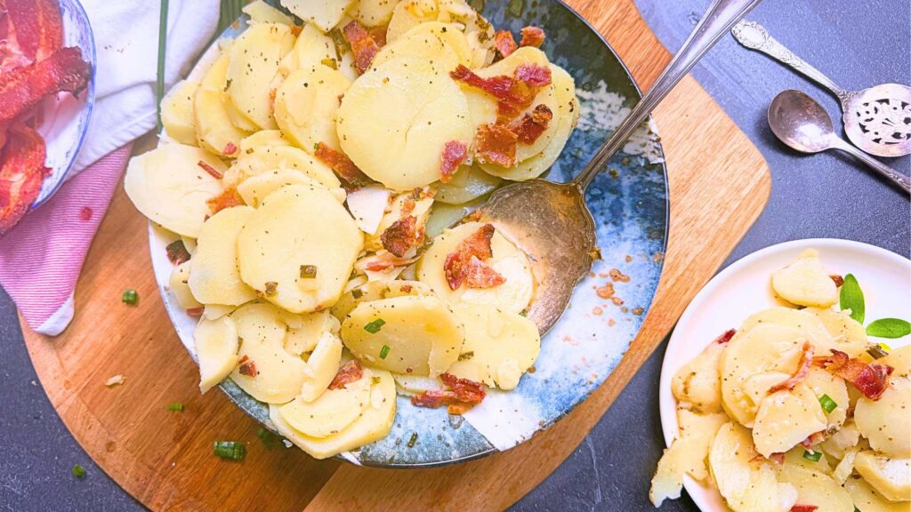 A large bowl of warm German potato salad with bacon. There is a serving on a small plate off to the side.