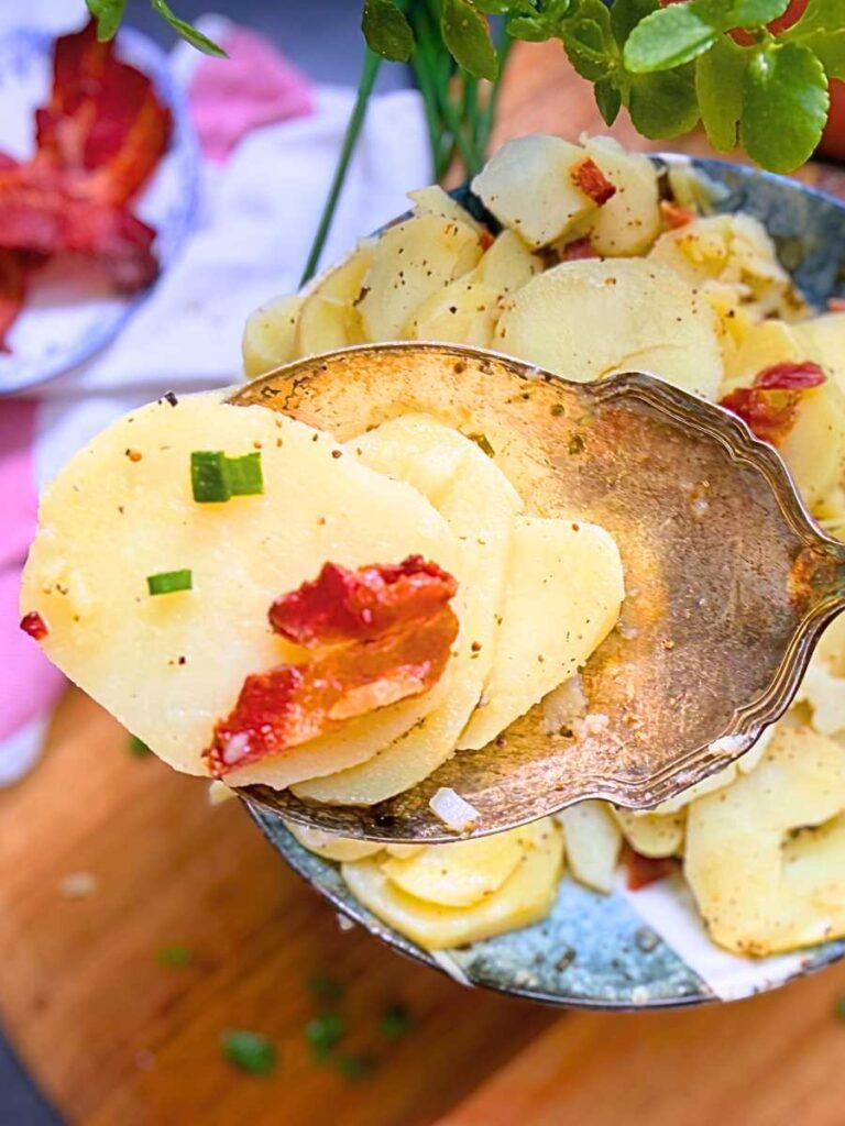 A large metal serving spoon holding some sliced potatoes with some bacon and chives.