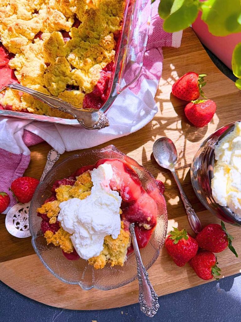 A baked strawberry cobbler in a square glass casserole dish. There is a serving of the strawberry dessert in front with whipped cream on top.