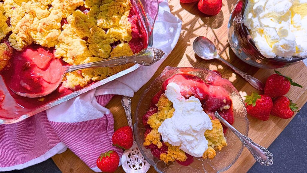 A baked strawberry dessert with a biscuit topping in a glass casserole dish. There is a serving in a glass bowl in front with whipped cream on top.