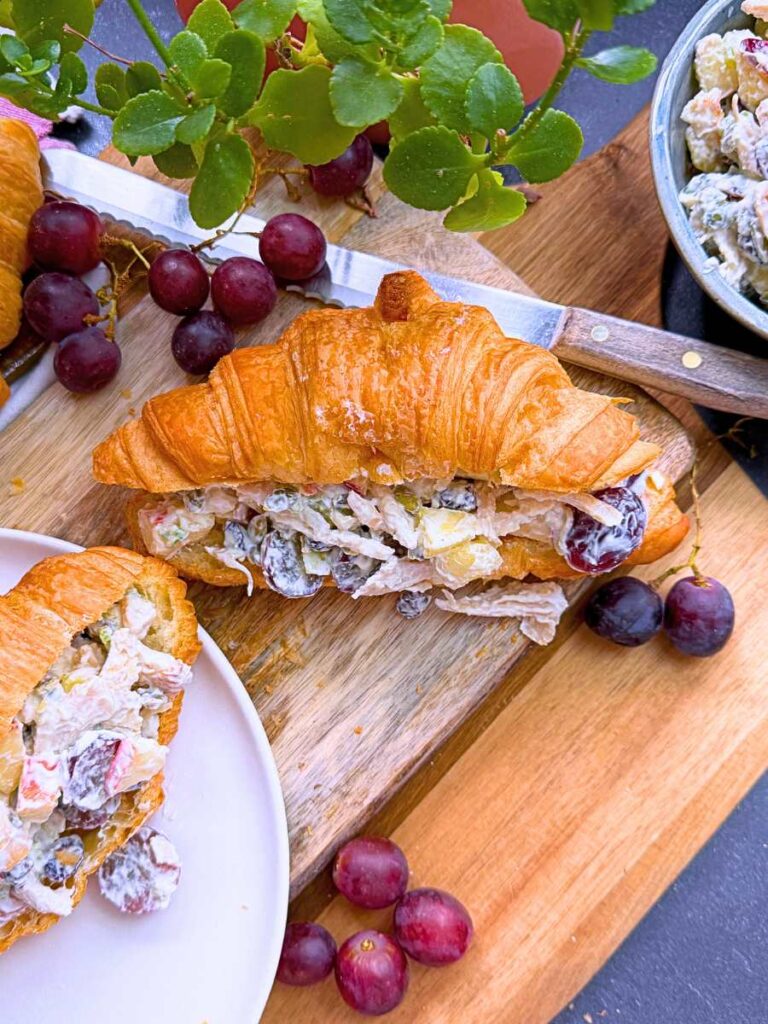 An overtop view of two croissant sandwiches filled with Waldorf chicken salad. There are purple grapes around the sandwiches.
