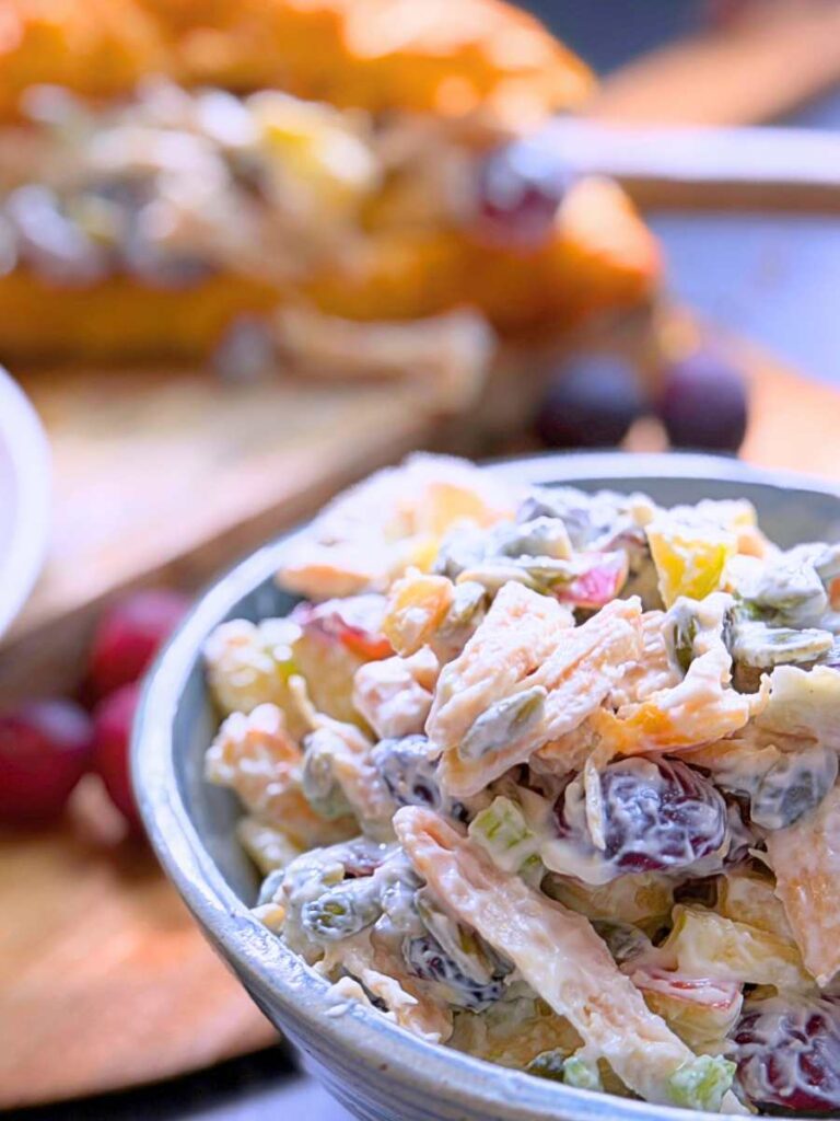 A bowl filled with Waldorf chicken salad. There is a croissant sandwich in the background.