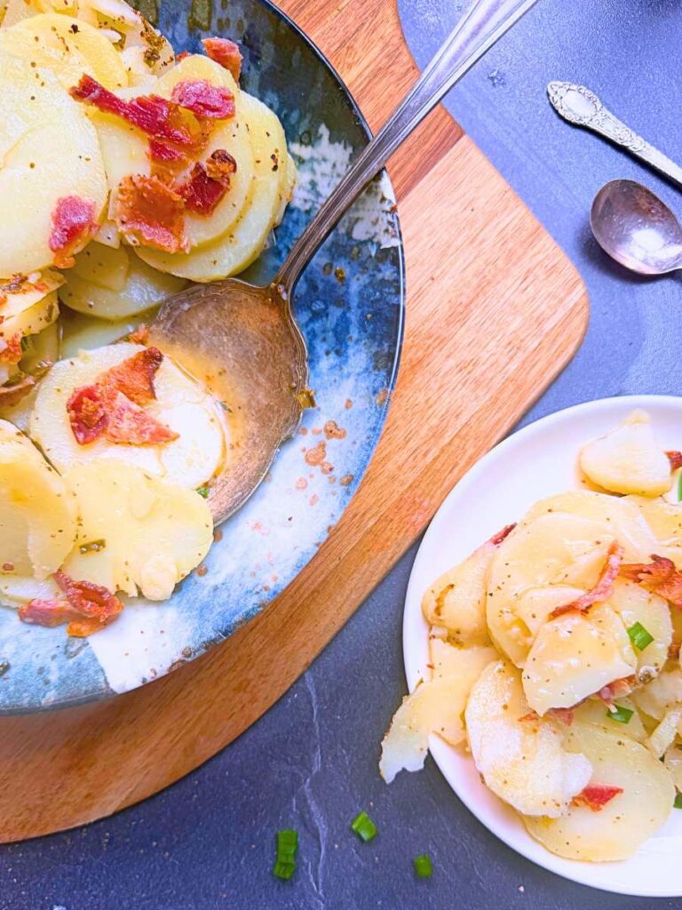 An overhead view of potato salad with sliced potatoes, bacon, and a vinegar dressing. There is a serving off to the side.