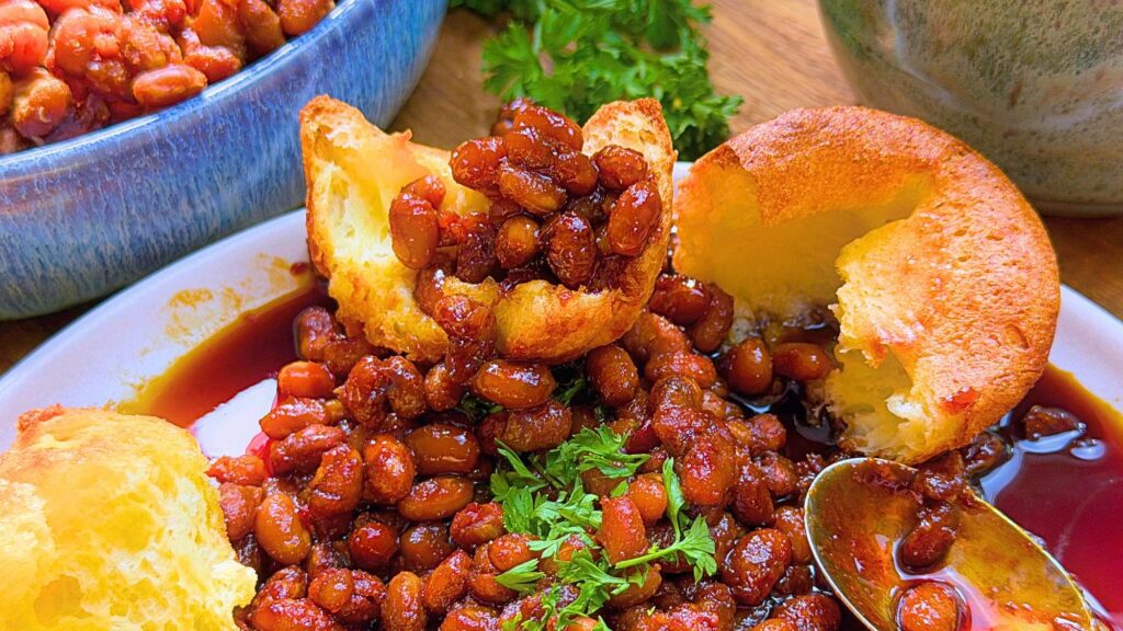 A plate of baked beans in a molasses sauce. There is a popover bun split in on the plate.