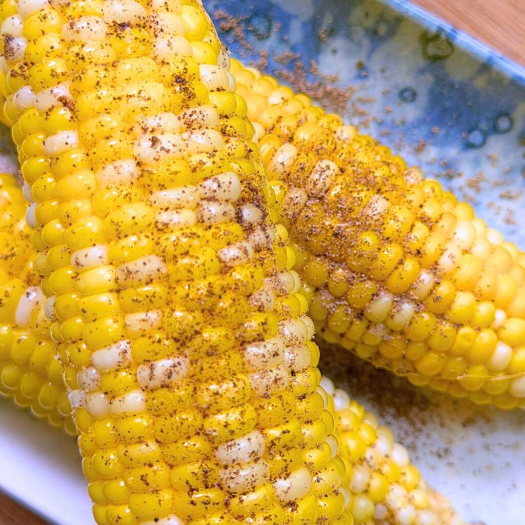 A close up look at three cooked ears of corn with melted butter, salt, and pepper. They are on a blue plate.