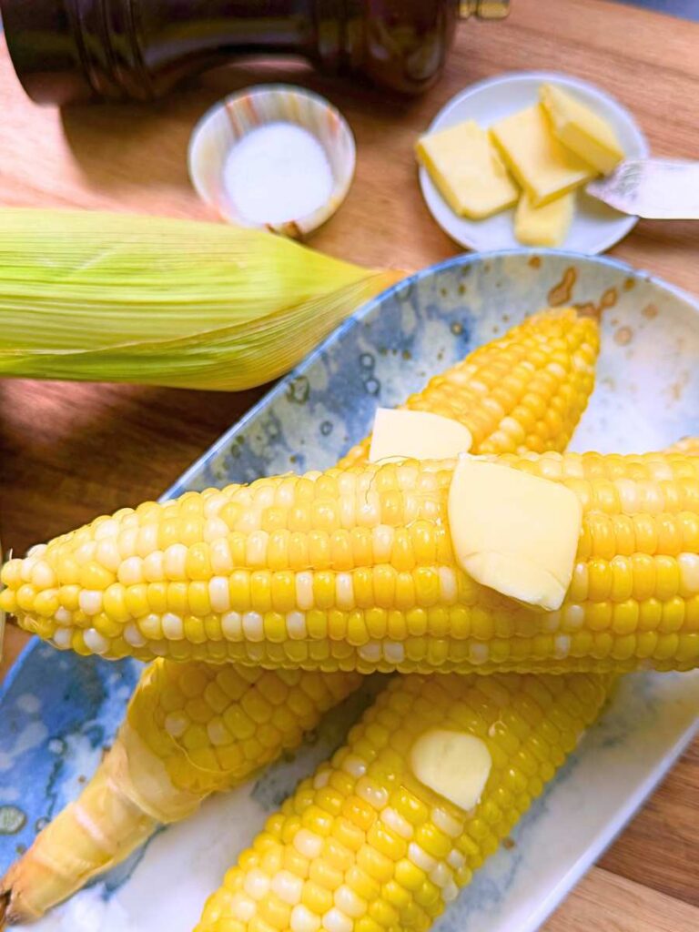 Three cooked ears of corn with pats of butter on top of each. There are some uncooked ears of corn off to the side.