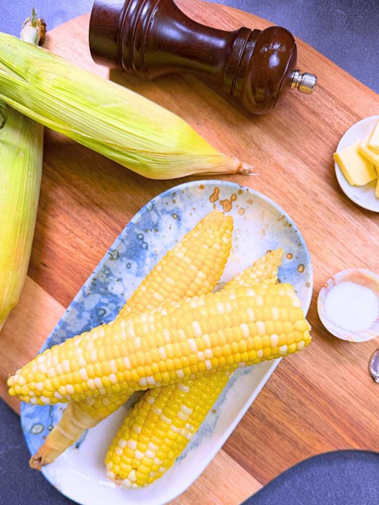 Three boiled ears of corn on a blue plate. There are some uncooked ears of corn with the husks on in the background.