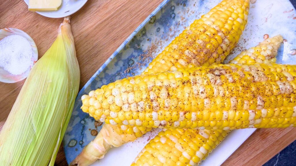 Three boiled corn on the cob with melted butter and salt and pepper. They are on a blue plate. There are some uncooked ears of corn off to the side.