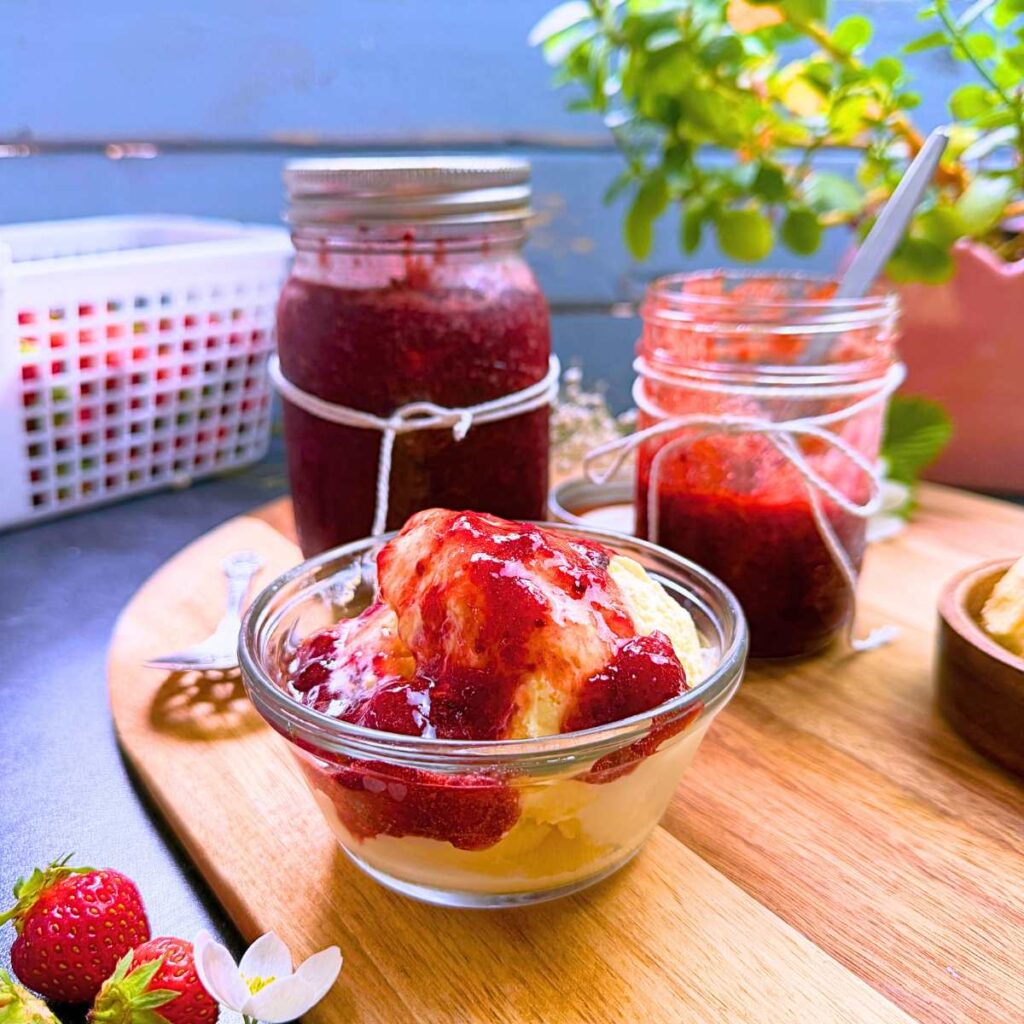 Two jars of strawberry jam sweetened with honey. One is opened and there is a wooden bowl of ice cream with strawberry jam on top.