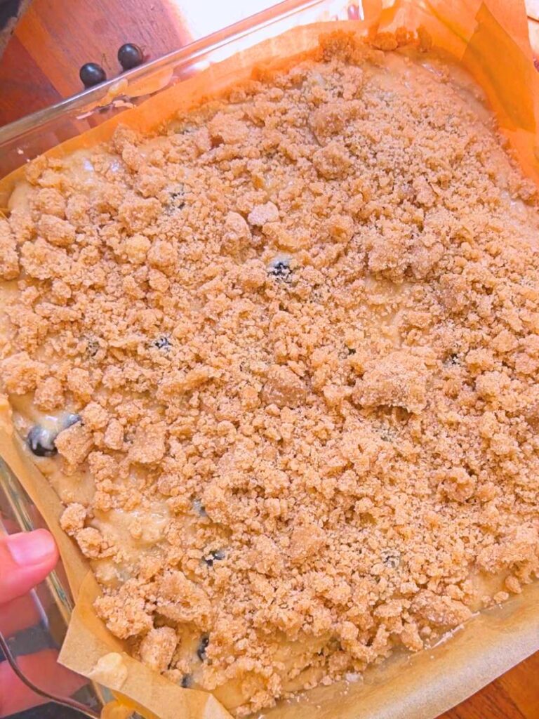 A black current coffee cake batter with a streusel topping in a lined, glass, square pan.