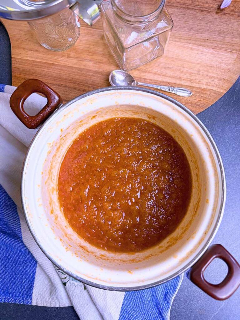 A white sauce pot with cooked mashed peaches inside. There are glass jars behind. There is a metal funnel on one jar.