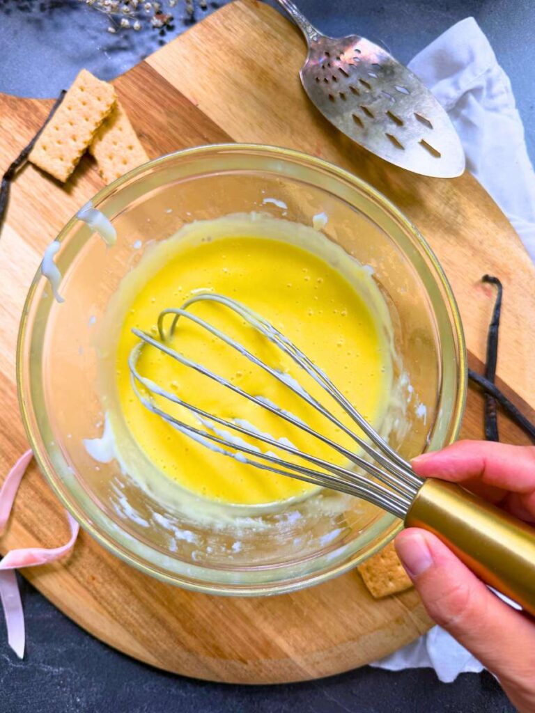 An egg yolk mixture in a glass bowl. A woman is holding a whisk.