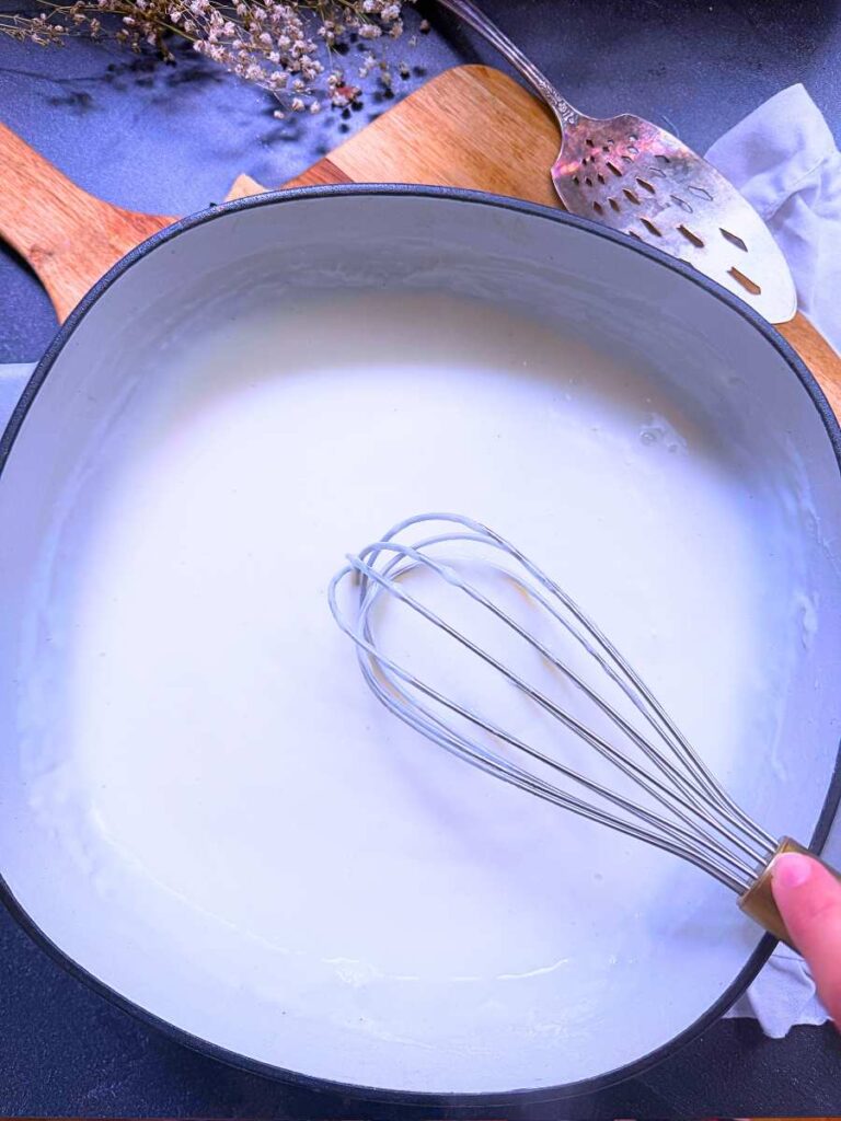 A large grey sauce pan with a milk mixture inside. A woman is holding a whisk.