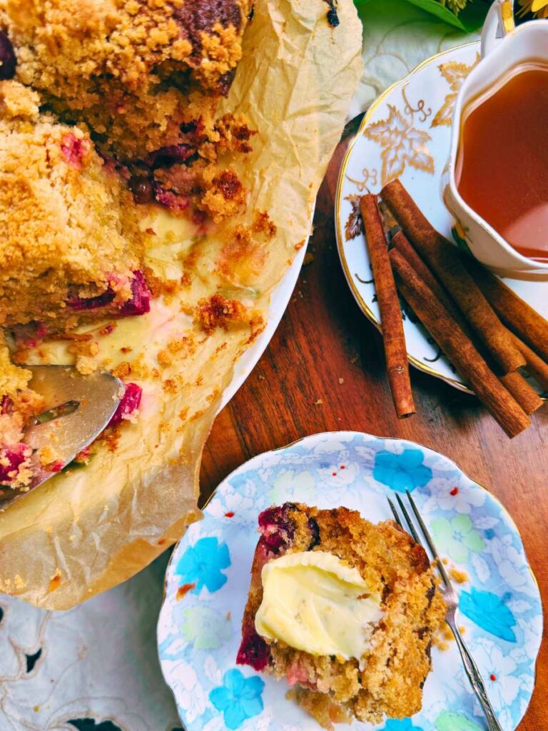A slice of gooseberry coffee cake with butter spread on top. There is more on a cake stand beside and tea with cinnamon sticks behind.