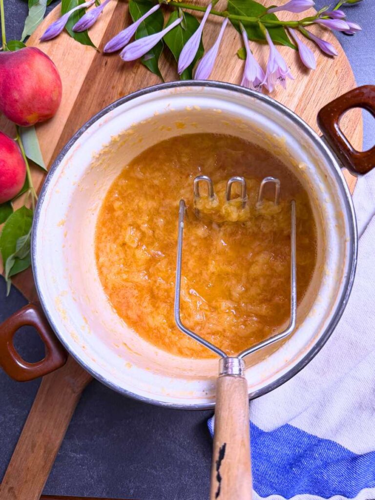 Mashed peaches in a white sauce pot. There is a metal masher with a wooden handle inside.