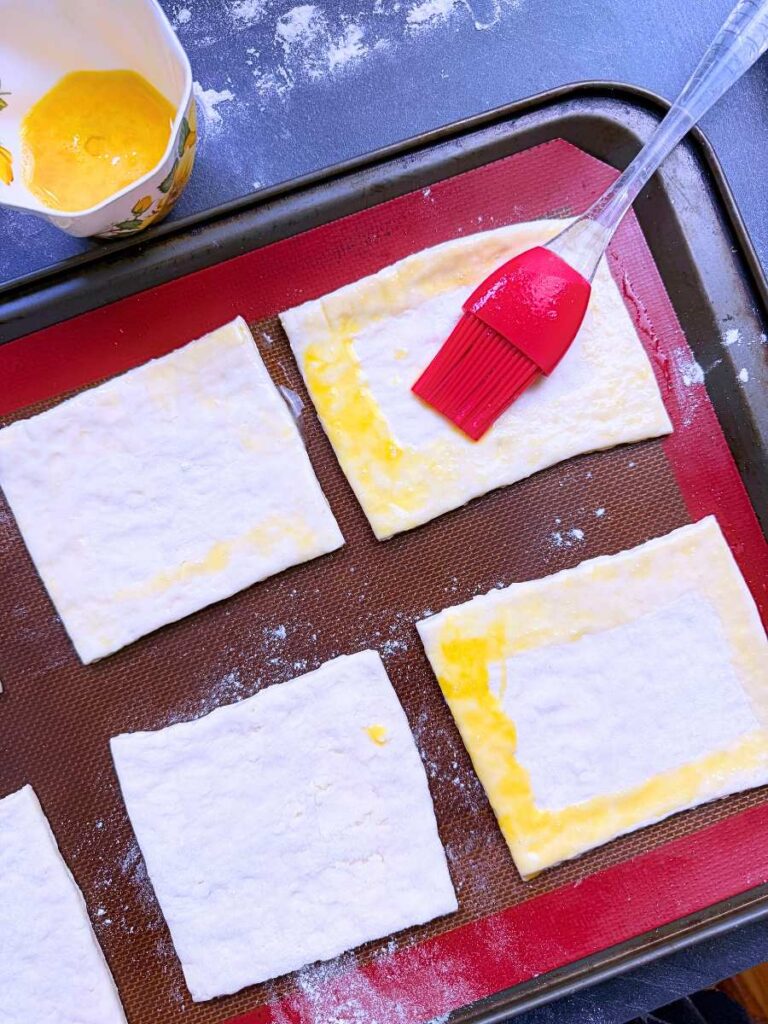 Squares of dough on a lined baking sheet. A woman is brushing an egg wash on to them.