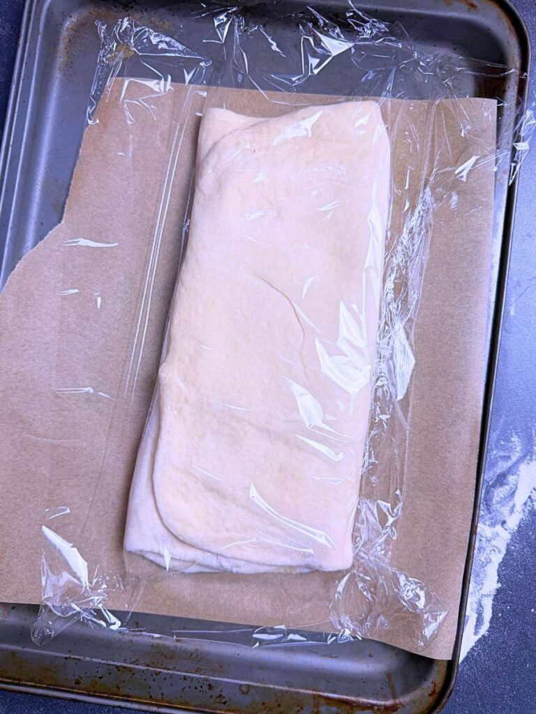 A baking sheet with folded dough covered in plastic wrap.