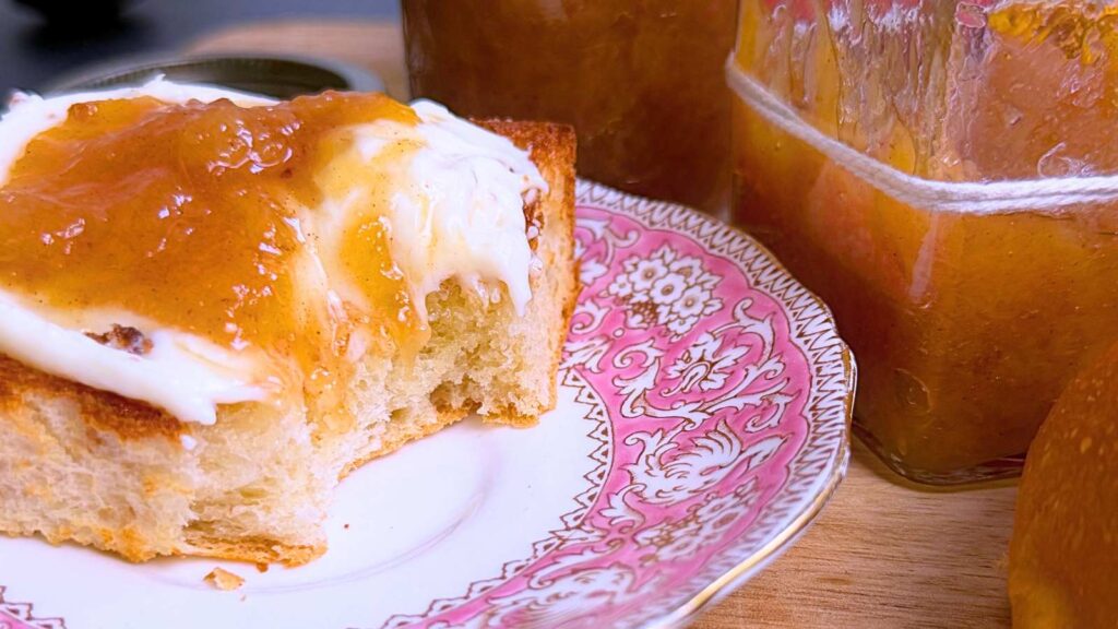 Half a bun spread with cream and peach jam on top. There are two jars of peach jam off to the side.