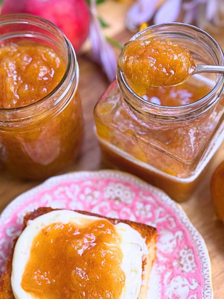 Two jars of peach jam. One has a spoonful resting on top. There is a bun on a pink plate spread with cream and peach jam.