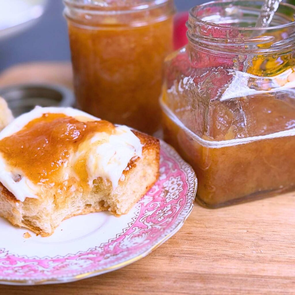 Half a bun with cream and peach jam on top. There is a bite out of the bun. There are two jars of peach jam in the background.