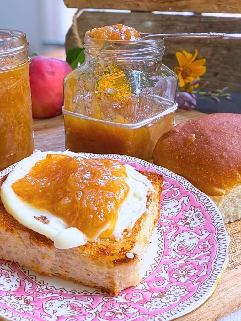 A toasted bun split in half on a pink plate. It is spread with cream and topped with peach jam. There are peach jam jars in the background.