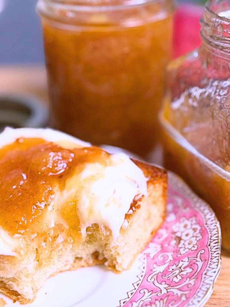 Two jars of peach jam. In front is a toasted bun spread with cream and topped with peach jam.