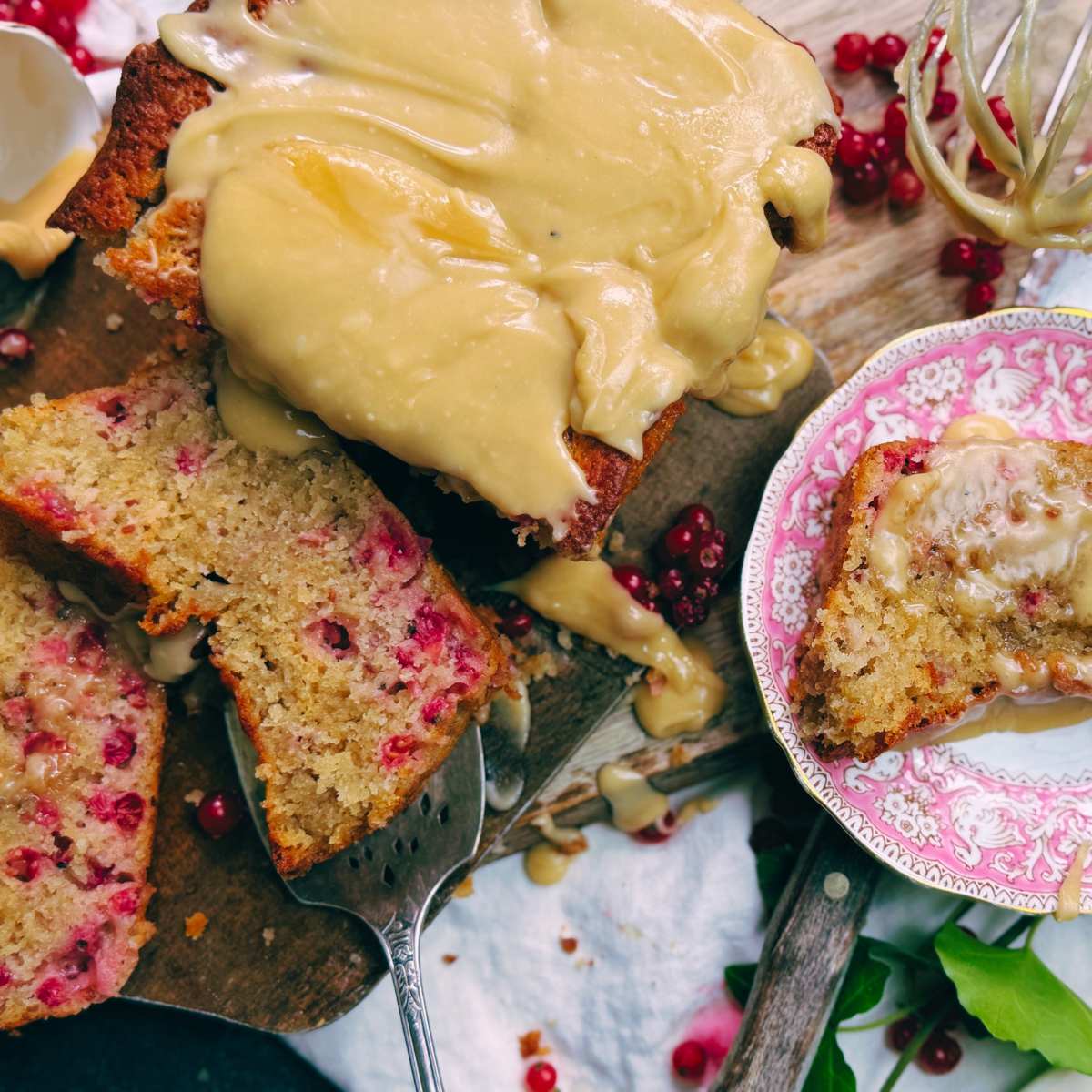 A sliced red current coffee cake with brown sugar icing. One slice is on a pink plate to the side.