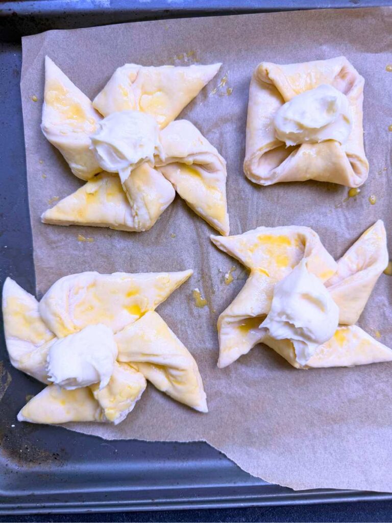 Unbaked danishes on a lined baking sheet. There is a cream cheese filling on top of each.