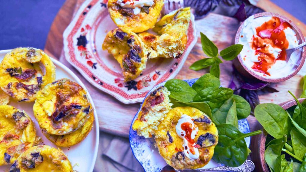 A plate full of tomato basil mini frittatas on a white plate. There are more off to the side on smaller plates and a bowl with cream and hot sauce.