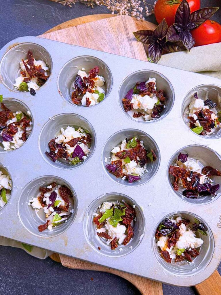 A muffin tin with chopped sun dried tomato, basil, and feta inside.