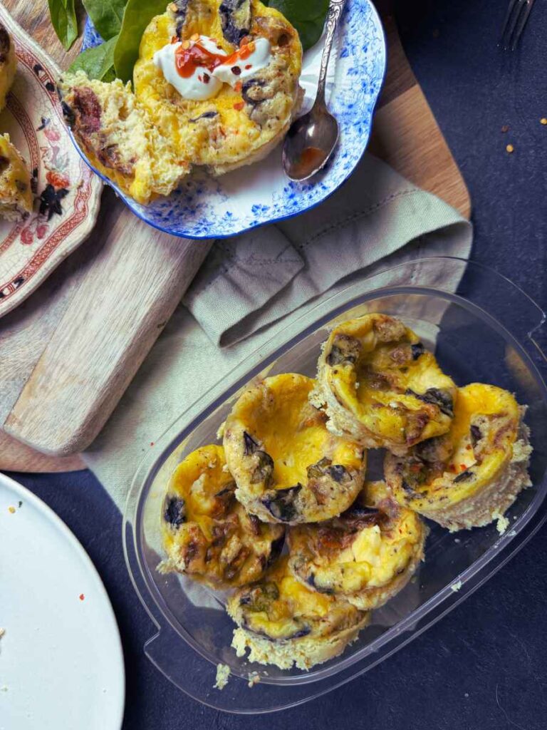 Muffin tin frittatas in a clear container. There is one on a blue plate in the background.