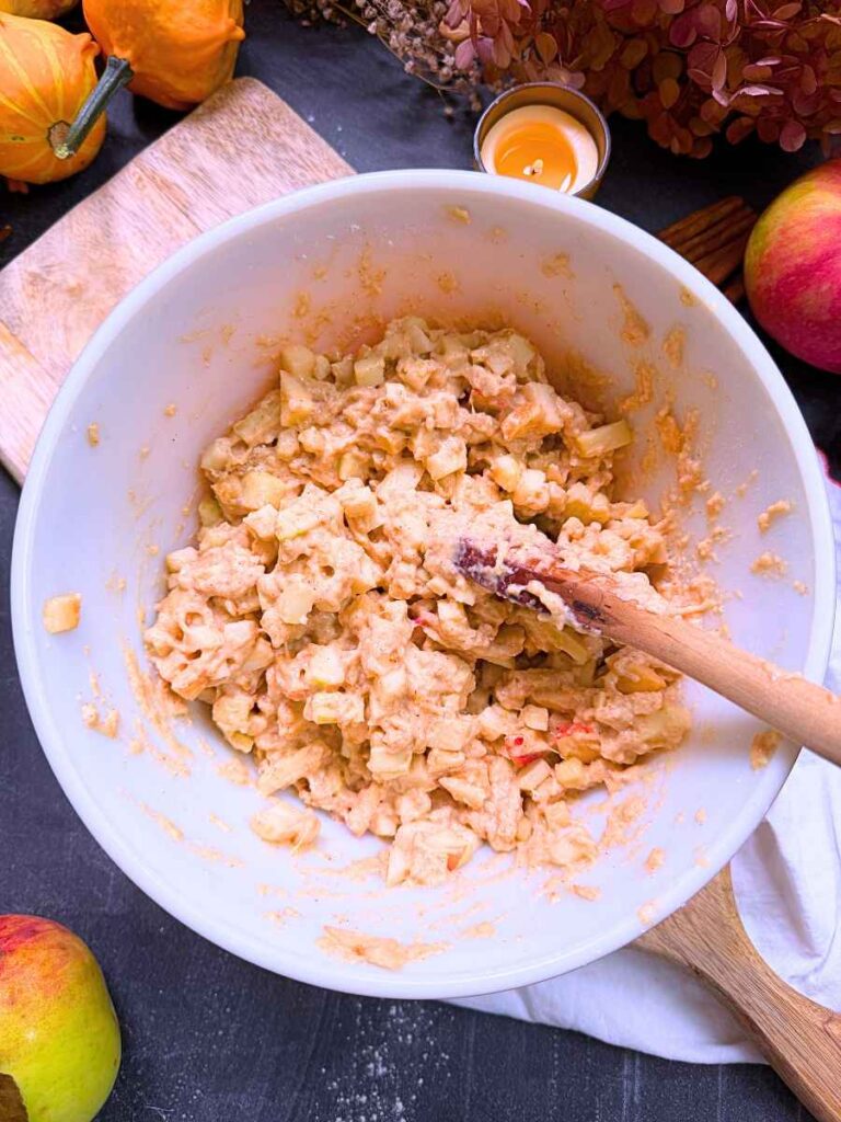 A white bowl with a apple fritter batter being mixed by a woman holding a wooden spoon.