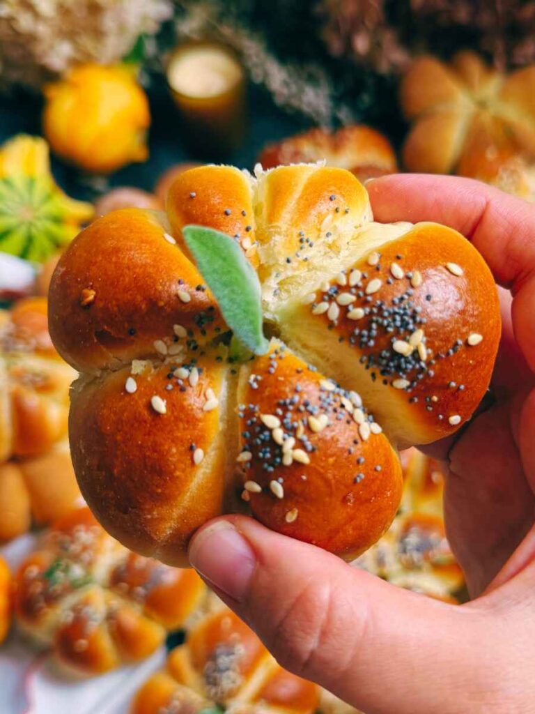 A woman is holding up a pumpkin-shaped dinner roll decorated with a sage leaf.