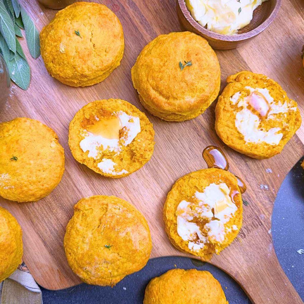 A wooden board lined with pumpkin biscuits. Some are split in half, buttered, with a drizzle of syrup.