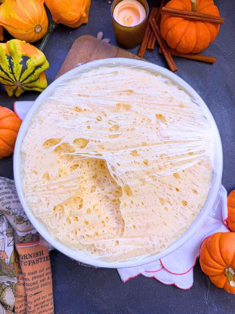 Risen dough in a white bowl covered with plastic wrap.