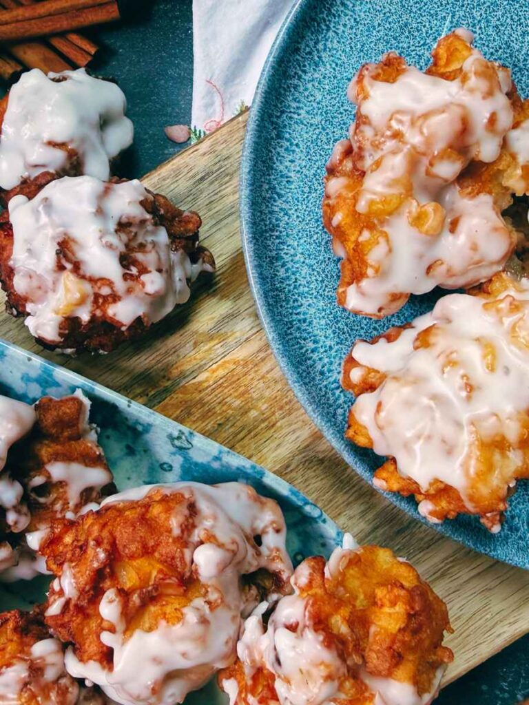 A collection of apple fritters. Some are on a blue plate, some on a white and blue plate, and some are off to the side on a cutting board.