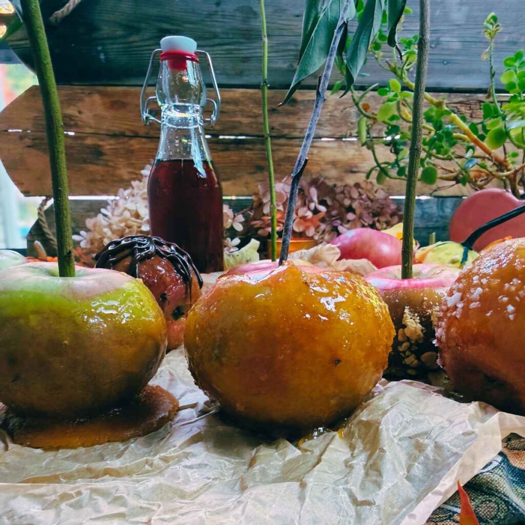 Two maple candy apples on a piece of parchment paper. There are more in the background as well as a bottle of maple syrup.