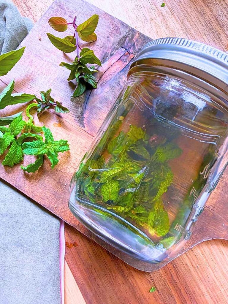 A glass jar with mint leaves and vodka inside. There are more fresh mint leaves off to the side.