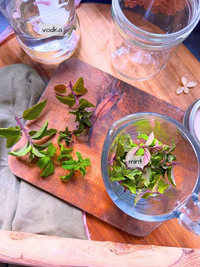 A wooden cutting board with a measuring cup of mint leaves and a bottle of vodka in the background.