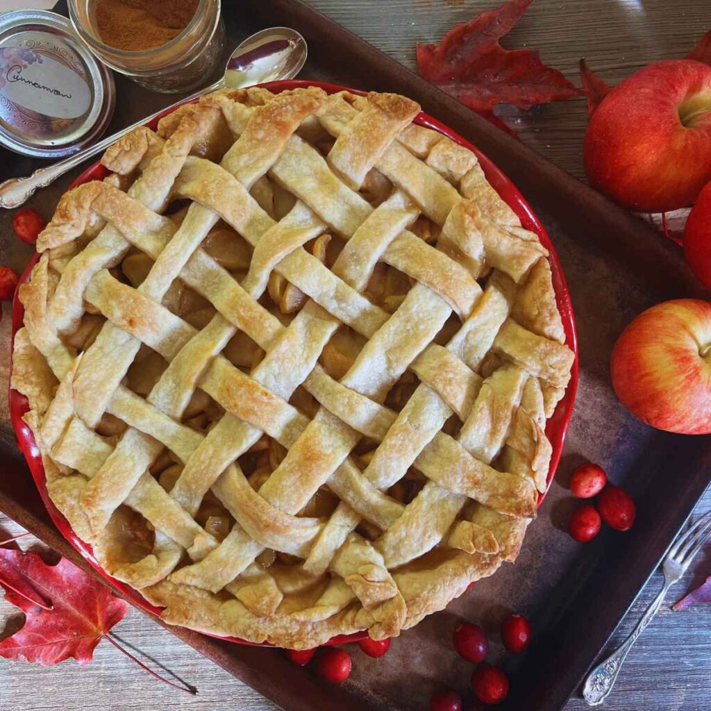 A lattice top apple pie with two whole apples off to the side and some fresh cranberries.