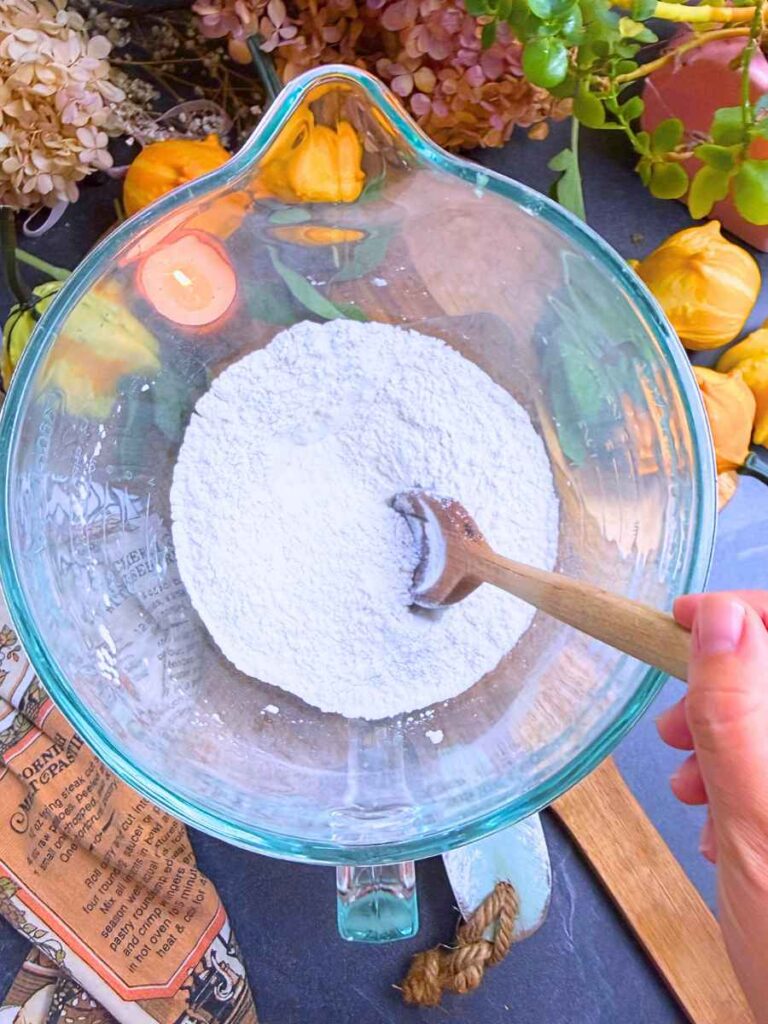 A large glass bowl with dry ingredients and a wooden spoon.