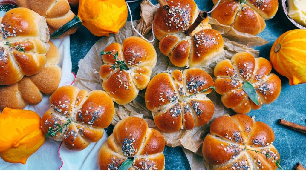 A collection of pumpkin shaped bread rolls. They have poppy and sesame seed tops and are decorated with fresh herbs and cinnamon sticks.