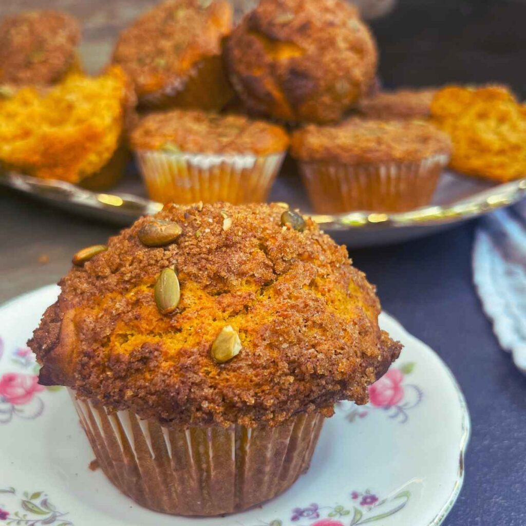 A pumpkin muffin topped with pumpkin seeds and a brown sugar crumble. There are more in the background.