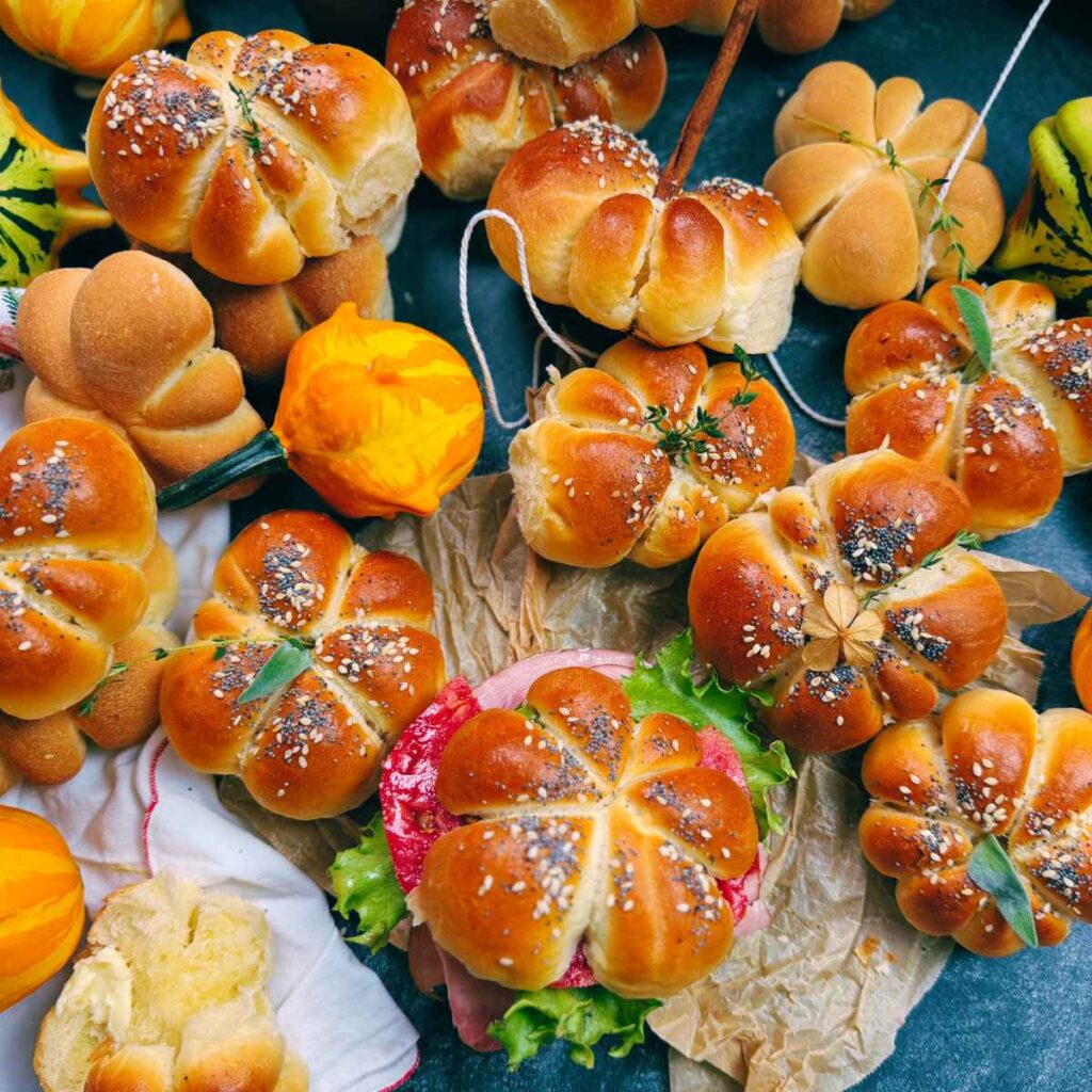 A collection of pumpkin-shaped dinner rolls on a table with fresh gourds around.