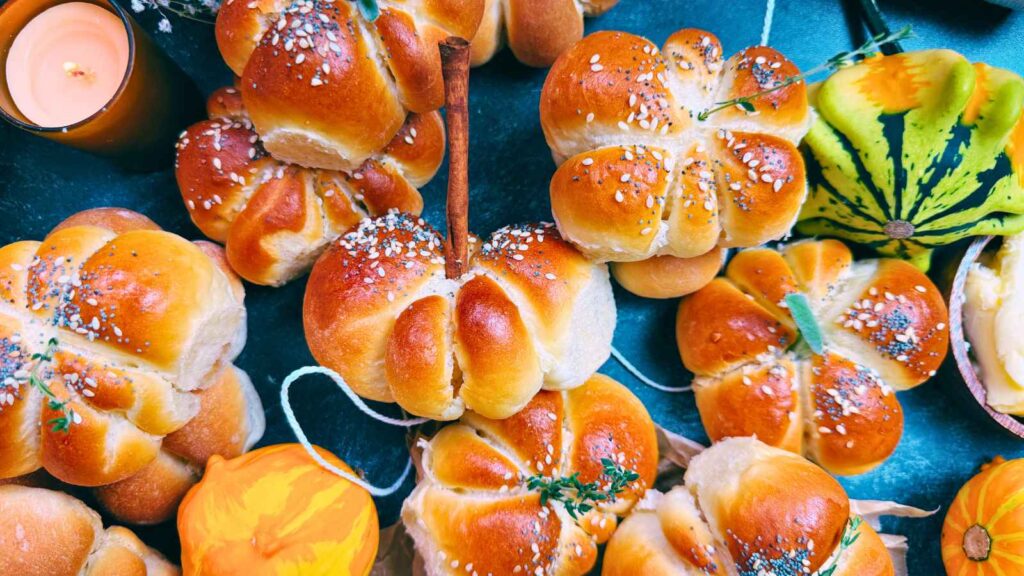 A collection of dinner rolls that are shaped like pumpkins are on a table some have fresh herbs and some have cinnamon sticks decorating them.