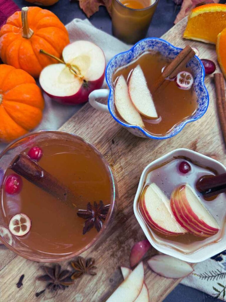 Three cups of mulled apple cider with sliced apples and whole spices. The background has mini pumpkins.