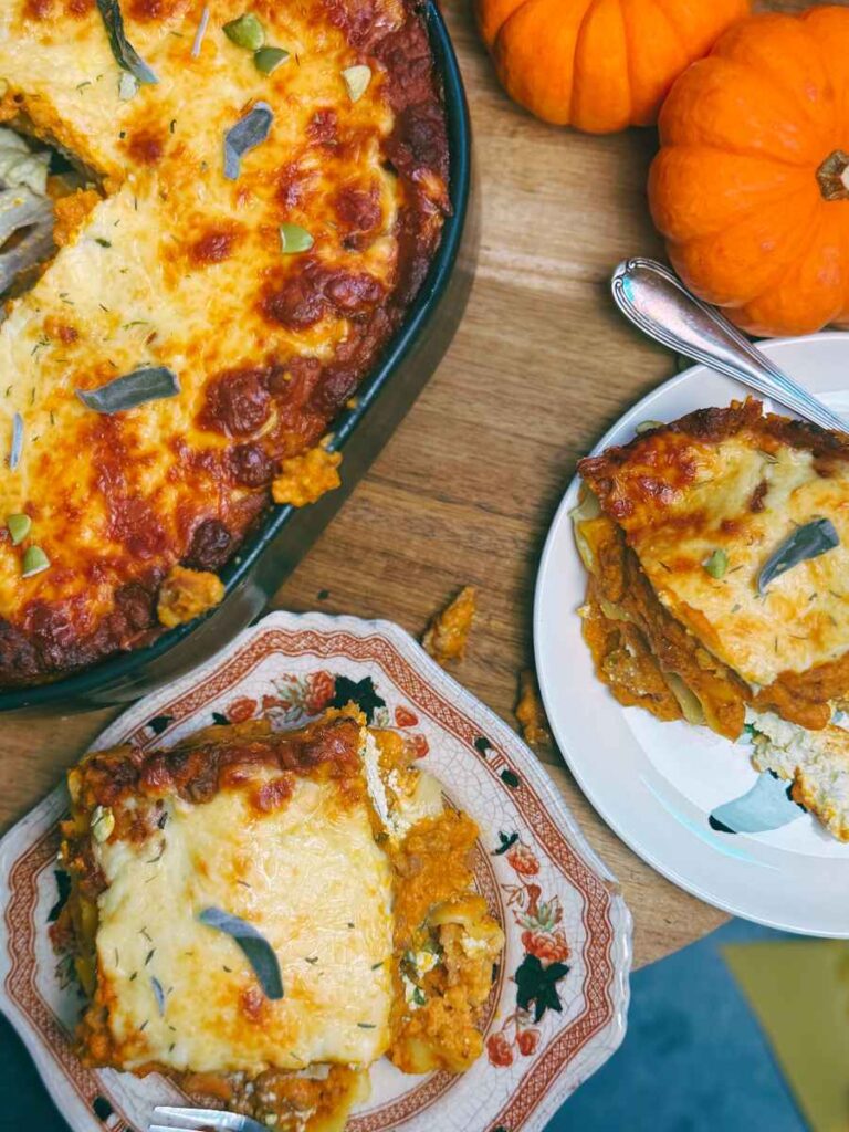 A baked pumpkin lasagna with two portions removed onto plates. There are sage leaves and pumpkin seeds on top.