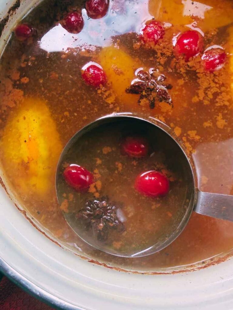 A white pot full of mulled apple cider with fresh fruit and whole spices. There is a scoop inside.